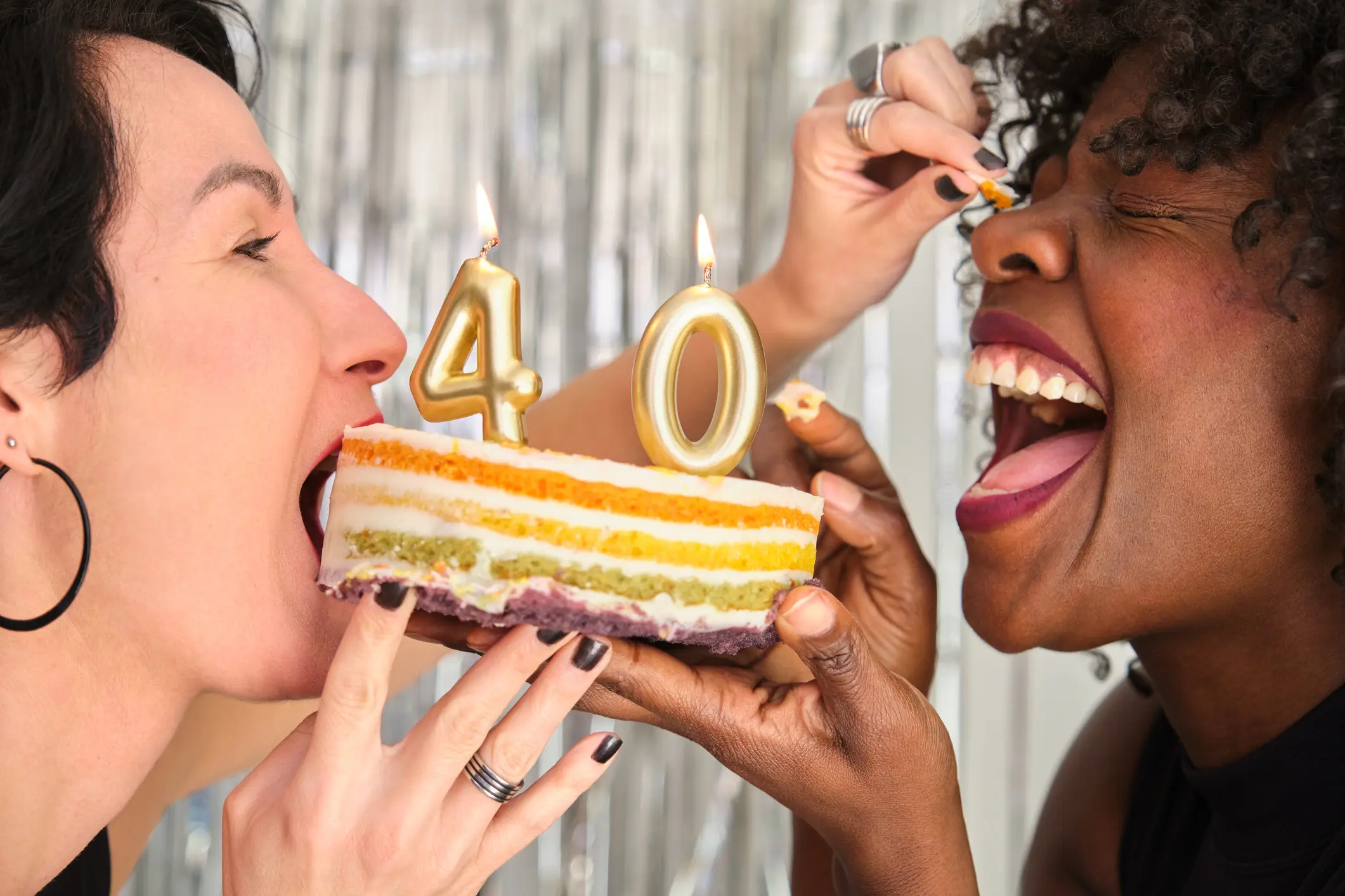 Two women holding a cake with lit 4 and 0 candles between them. The one with the cake in her mouth is tryng to put cake on the other's nose