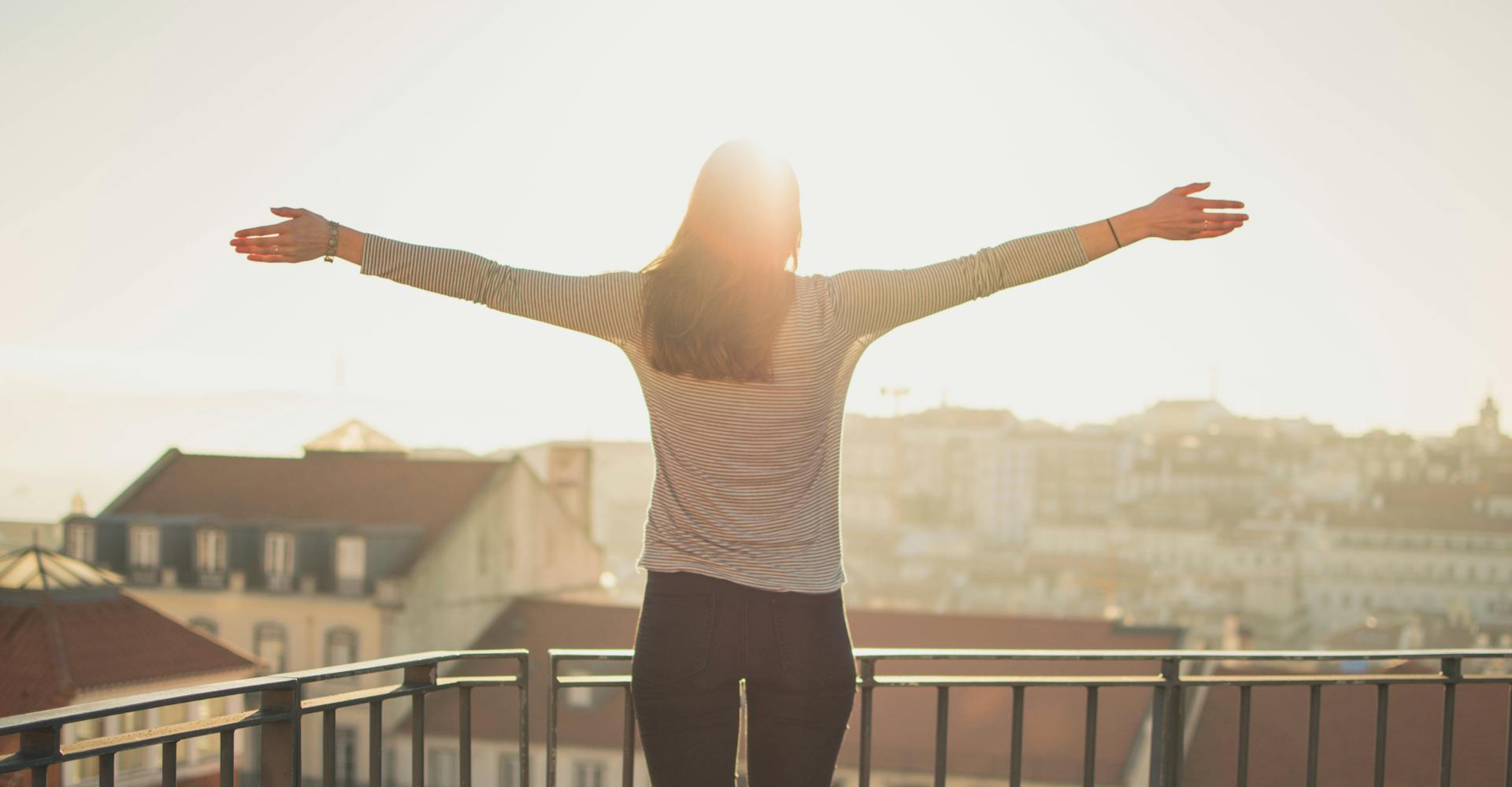 Woman with outstretched arms standing on her balcony facing the sunrise over the city
